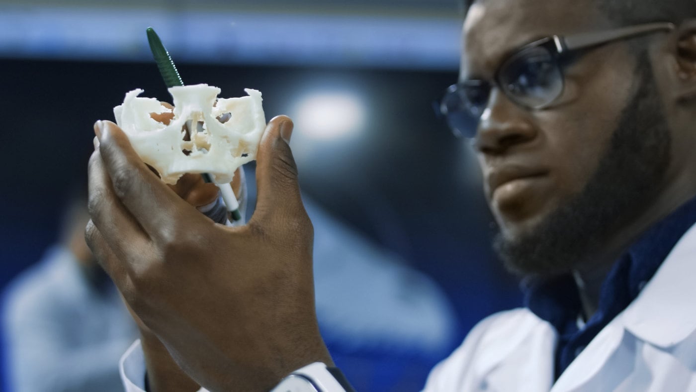 A scientist in glasses and lab coat closely examining a 3D-printed white object in a laboratory setting.