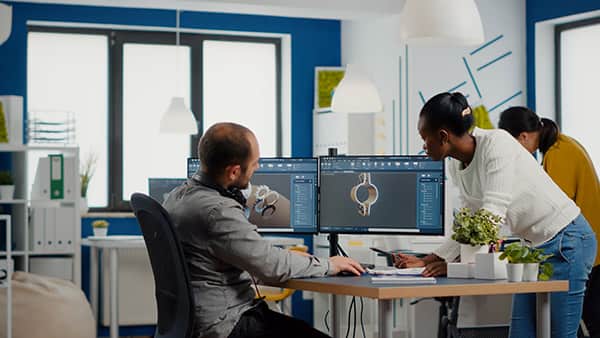 Two professionals, a man and a woman, collaborating over a computer in a modern office workspace. The screen displays graphic design software.