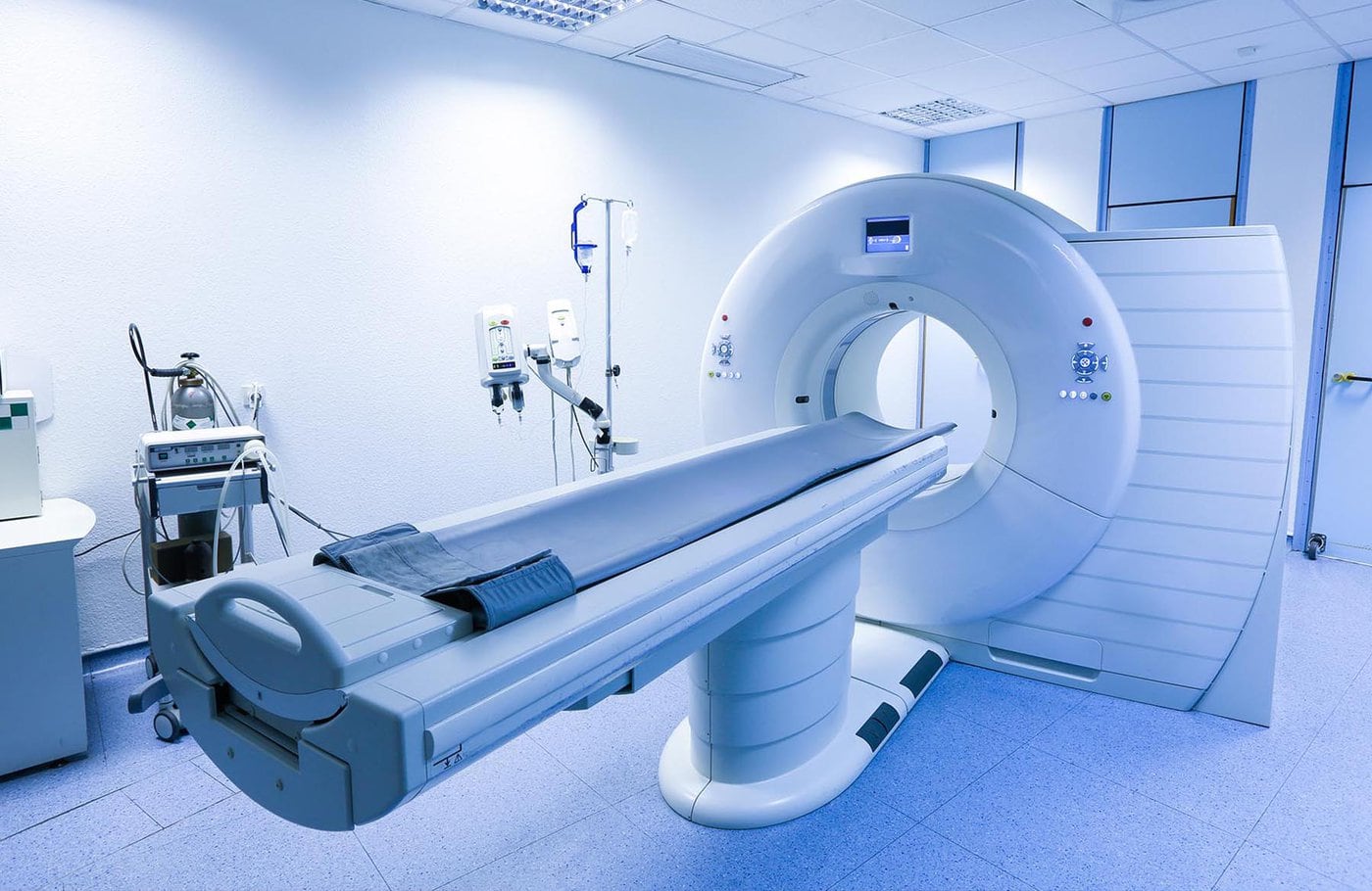 A Modern Mri Machine In A Clean, Well-Equipped Medical Imaging Room With White Walls And Various Medical Devices Around.