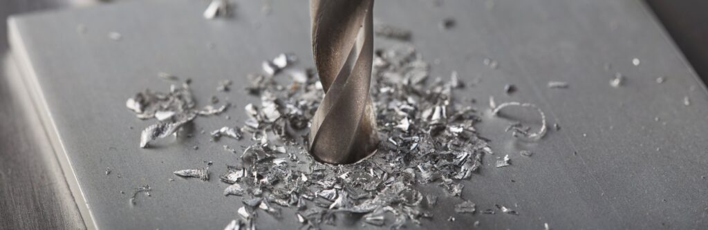 Close-Up Of A Metal Drill Bit Creating A Hole In A Flat Metal Surface. Metal Shavings Surround The Bit, Indicating Drilling Action. The Surface And Drill Bit Are Both Metallic And Silver In Color.