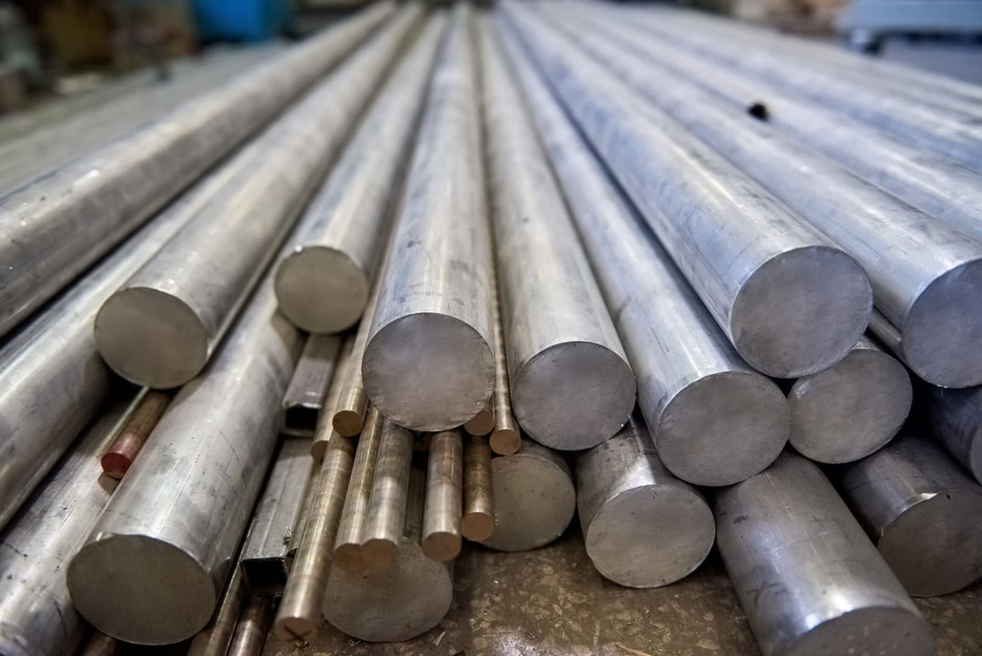 A collection of stacked metal rods in various sizes lies on a warehouse floor. The rods are shiny and cylindrical, reflecting industrial lighting, and some have a copper hue, suggesting different metal types.