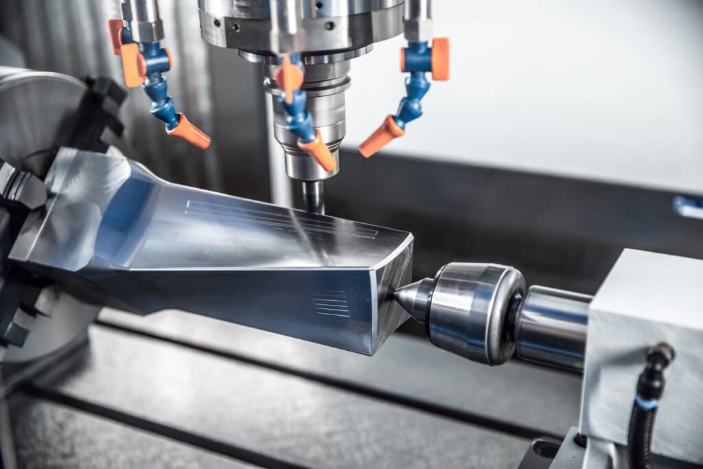 A Close-Up Of A Cnc Machine In Operation, With A Metal Piece Being Precisely Milled By A Spinning Tool. Multi-Directional Blue And Orange Nozzles Are Visible, Likely Used For Cooling Or Lubrication During The Machining Process.