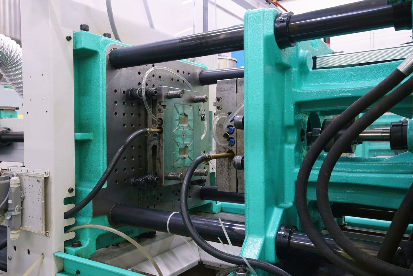 Close-up of a green and silver injection molding machine in a factory setting. The image shows the mold, with rods and cables connected, highlighting the industrial manufacturing process.