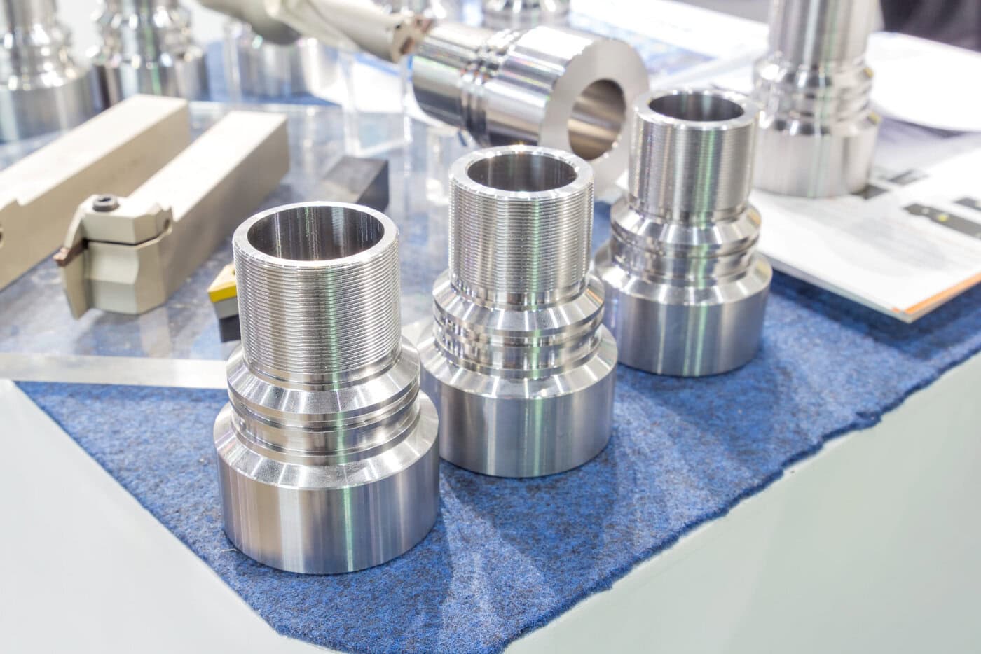 Three shiny, metallic cylindrical machine parts, crafted with precision through CNC machining services, are displayed on a blue fabric-lined table. They have threaded sections and are positioned in a row. Various other metal components are visible in the background on a reflective surface.