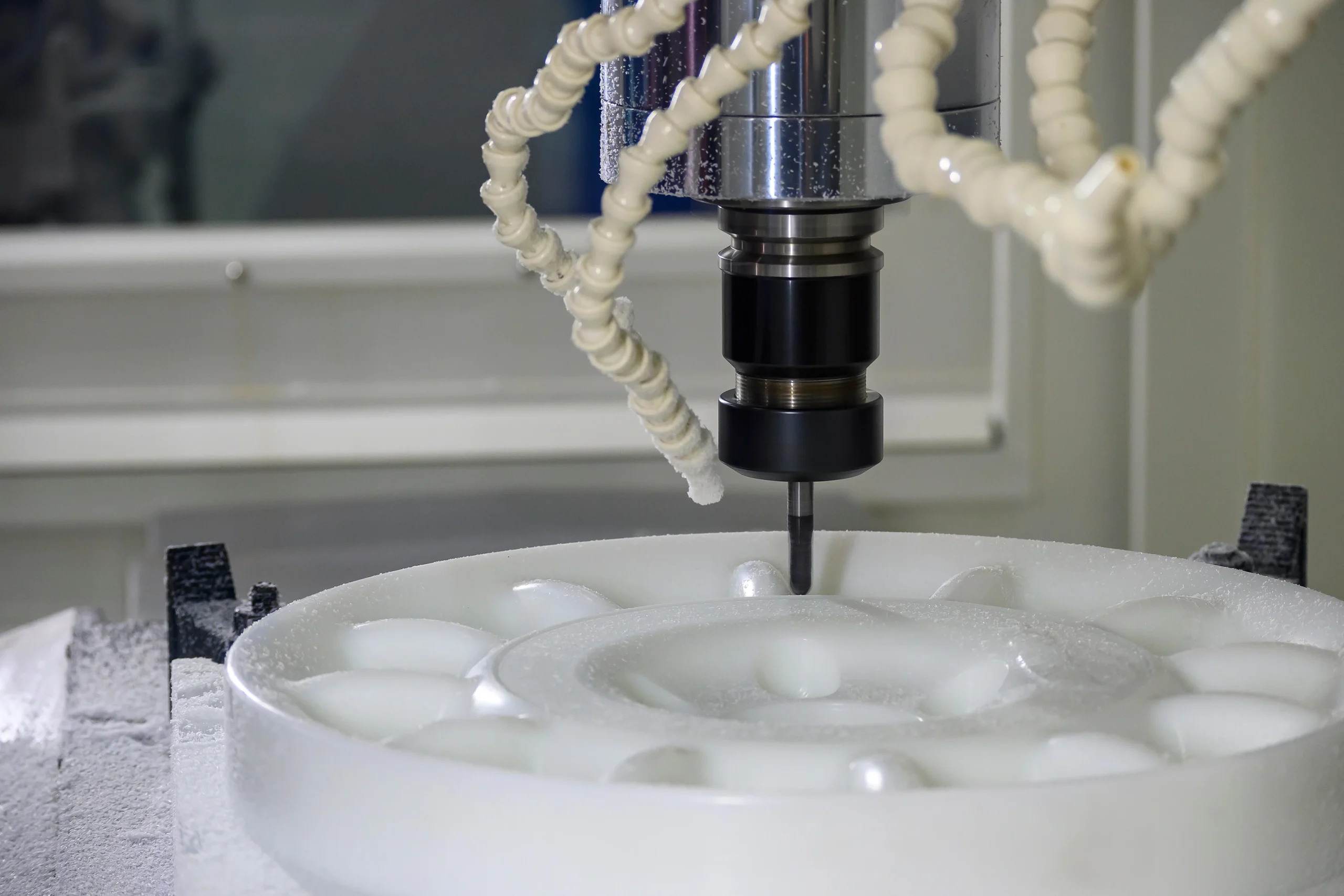 A CNC milling machine is carving a circular pattern into a white material. The machine's spindle and cutting tool are visible, surrounded by flexible coolant hoses. The setting suggests a precise industrial machining process.