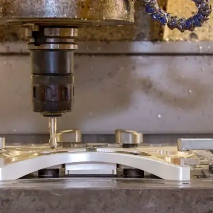 Close-Up Of A Cnc Milling Machine In Operation. The Machine'S Drill Bit Is Cutting Into A Metallic Piece Fixed In Place, Creating Precise Shapes And Designs. Metal Shavings Are Visible Around The Work Area, Indicating Active Machining.