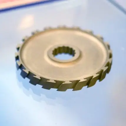 A Close-Up Photo Of A Metallic, Circular Gear With Evenly Spaced Teeth Around Its Edge, Placed On A Smooth, Reflective Surface. Created Using Dmls 3D Printing Service, The Gear Has A Central Hole With Ridged Inner Edges, Suggesting It Is Part Of A Mechanical Assembly.