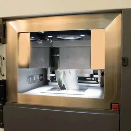 A 3D Printer In Operation, Encased In A Metallic Frame. The Dmls 3D Printing Service'S Extruder Is Actively Working On A Partially Completed Object Inside The Machine. The Object Is Illuminated By A Light Source From Within The Printer’s Enclosure.