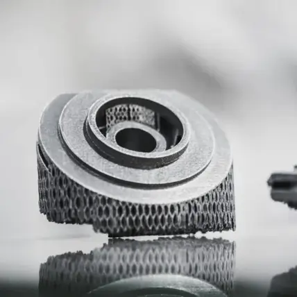 A Close-Up Image Of A 3D-Printed, Metal Mechanical Component On A Reflective Surface. The Component, Produced Using A Dmls 3D Printing Service, Features Intricate, Honeycomb-Like Internal Structures And Smooth, Circular Outer Layers. The Background Is Blurred, Emphasizing The Details Of The Object.