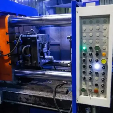 Image Of An Industrial Machine Used For Manufacturing, With Orange And Blue Components. The Machine, Ideal For Rapid Die Casting, Is Equipped With A Control Panel Featuring Numerous Buttons And Indicators. Metal Parts And Wires Are Visible, Along With A Workpiece In Progress.