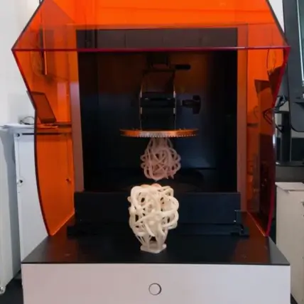 A 3D Printer In Operation, Housed Within An Orange-Tinted Enclosure, Is Creating A Complex, Interwoven Geometric Structure Using Sla 3D Printing Technology. Another Similar Finished Structure Rests On The Surface In Front Of The Printer.