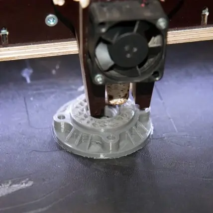 A 3D Printer Utilizing Fdm 3D Printing Technology Is In The Process Of Creating A Circular, Intricate Object. The Extruder Head, Equipped With A Small Cooling Fan, Is Depositing Filament Layers Onto The Build Platform. The Object Appears Partially Complete, With Fine Details Visible.