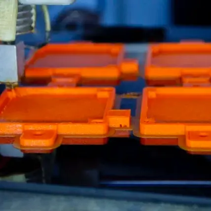 A Close-Up Of A 3D Printer Nozzle Depositing Layers Of Orange Filament Through Fdm 3D Printing To Create Four Rectangular Objects With A Small Tab. The Partially Finished Items Rest On The Printer Bed, While The Background Is Beautifully Blurred.
