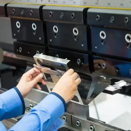 A Person In A Blue Shirt Is Operating A Hydraulic Press Brake Machine, Carefully Positioning A Prototype Sheet Metal Between The Machine'S Clamping System And Bending Tool. The Machine Has Gear-Like Components And Measurement Markings On Its Top Section.