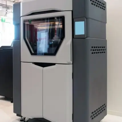A Sleek, Large Industrial 3D Printer, Designed For Fdm 3D Printing, Is Prominently Placed In A Well-Lit Room. The Machine Features A Gray And Black Color Scheme With A Transparent Front Panel, Revealing Some Of The Internal Components. Various Other Machinery Can Be Seen In The Background.