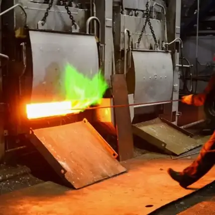 A Worker Is Holding A Rod With A Bright, Glowing Hot End, Emitting A Green Flame, In Front Of Large Industrial Furnaces. The Worker Is Wearing Safety Gear While Handling The Heated Material In An Industrial Setting.