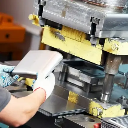 A Person Wearing Gloves Carefully Places A Prototype Sheet Metal Into An Industrial Stamping Machine. The Machine, With Its Yellow Painted Base, Stands Ready For Action. Nearby, Multiple Stacked Metal Sheets Await Their Turn.