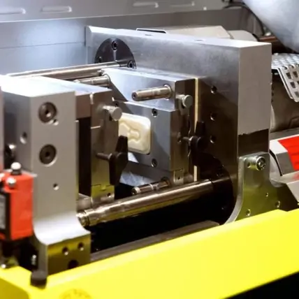 A Close-Up Of A Prototype Injection Molding Machine In Operation. The Machine Has Several Metal Components And A Mold Cavity Where Plastic Material Is Being Shaped. The Setup Is Inside A Metallic Framework, And Part Of The Yellow Machine Base Is Visible.