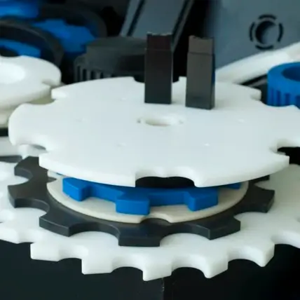 Close-Up Of Interlocking Plastic Gears In Black, White, And Blue. The Prototype Injection Molding Gears, Assembled Together, Showcase Their Various Sizes And Teeth, Forming A Complex System. The Background Is Blurred, Emphasizing The Detailed Structure Of The Gears In The Foreground.