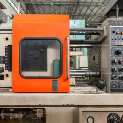 A Large Prototype Injection Molding Machine With An Orange Safety Enclosure On The Left Side. The Enclosure Has A Small Clear Window And A Handle. On The Right, There Is A Control Panel With Numerous Buttons, Switches, And Dials. The Machine Is Set In A Factory Environment.