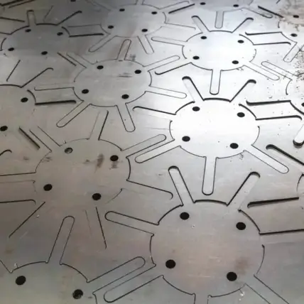 A Prototype Sheet Of Metal With Intricate Geometric Cutouts, Created Through Laser Cutting, Forms A Repeating Pattern Of Circular Shapes With Extended Lines. The Metal Has A Slightly Worn And Industrial Appearance, With Some Small Spots And Markings Visible On The Surface.