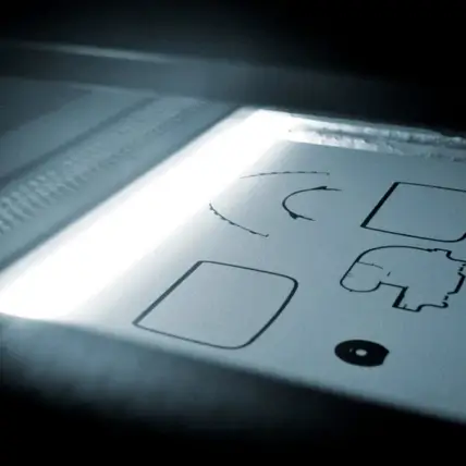 A Close-Up Image Showing A Sheet Of Paper Being Scanned Under A Bright Light. The Paper Has Several Shapes, Including Squares, A Crescent, And An Abstract Figure, Along With Some Text Hinting At Mjf 3D Printing Services. The Scene Is Dimly Lit Except For The Scanner'S Light.