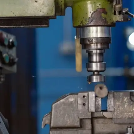 A Close-Up Image Of A Milling Machine In A Workshop. The Machine Head Is Green And Is Positioned Above A Metal Workpiece, Ready For Machining. The Background Is Blurred, Featuring Out-Of-Focus Machinery And A Blue Wall.
