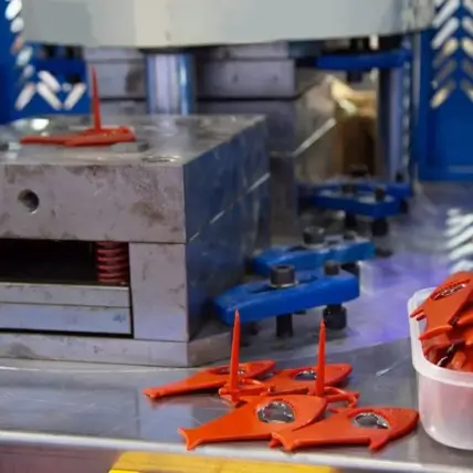 The Industrial Machine Hums In Operation, Expertly Handling Prototype Injection Molding. Red Plastic Parts With Pointed Ends Are Manufactured And Scattered Around The Machine, Then Collected In A White Container. The Background Reveals An Array Of Machinery Components And Metal Parts At Work.