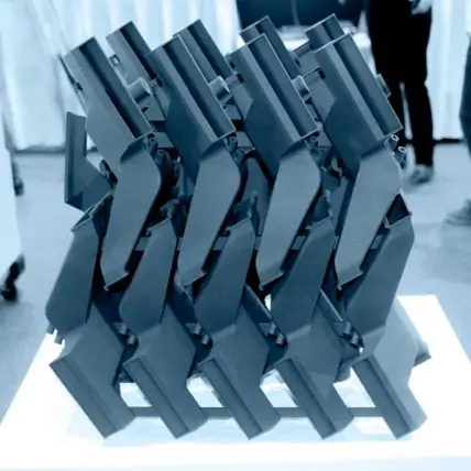 A Modern Art Installation Features Multiple Black Handguns Intricately Arranged In A Geometric, Lattice-Like Pattern, Seemingly Crafted With Precision Akin To Mjf 3D Printing Service. The White Pedestal Anchors The Display, While The Blurred Background Reveals Indistinct Human Figures And White Curtains.