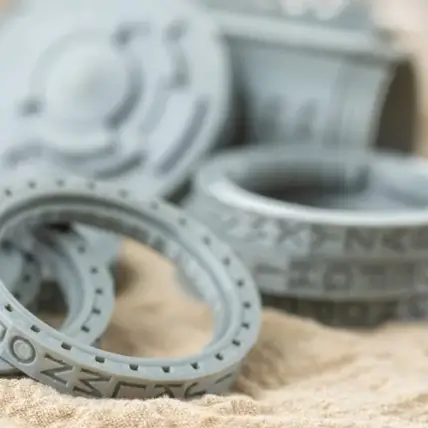 A Close-Up Of Several 3D-Printed, Round Objects With Intricate Designs And Patterns, Crafted Using A Polyjet 3D Printing Service. The Objects Are In Various Shades Of Gray And Appear To Be Made From A Plastic-Like Material. They Rest On A Beige, Textured Surface.