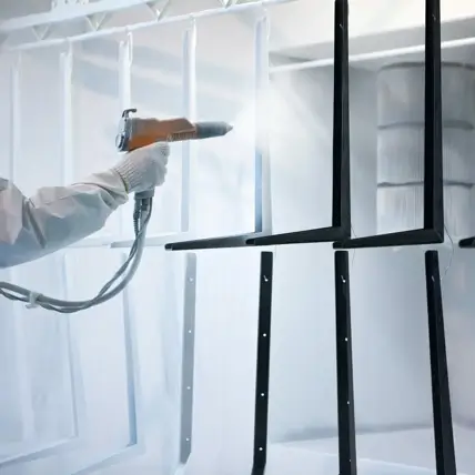 A Person Wearing Protective Clothing And Gloves Uses A Spray Gun To Apply White Powder Coating To Metal Frames Hanging From A Rack In A Workshop Or Industrial Setting.