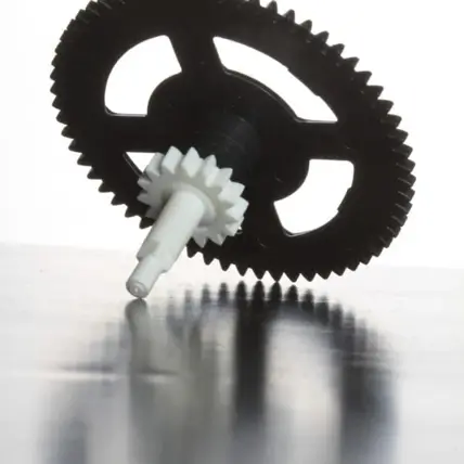 Close-Up View Of A Black Gear, Created Using An Sls 3D Printing Service, With A Central White Spindle Partially Reflected On A Shiny Surface. The Gear Has A Circular Form With Evenly Spaced Teeth Along The Outer Edge And Three Spokes Connecting To The Central Hub. The Background Is White And Minimalistic.