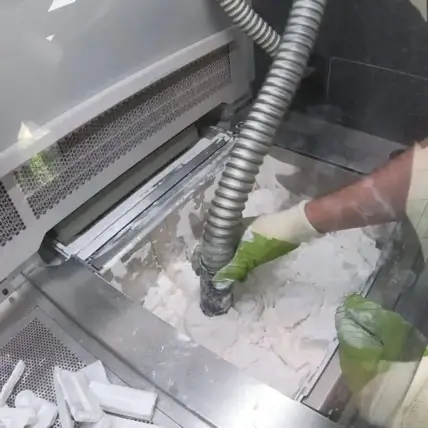A Person Wearing Green Gloves Is Operating A Piece Of Machinery, Using A Large Vacuum Hose To Suck Up Fine White Powdery Material. The Work Area Appears Industrial, With Metallic Surfaces And A Mesh Ventilation System, Likely Part Of An Sls 3D Printing Service.