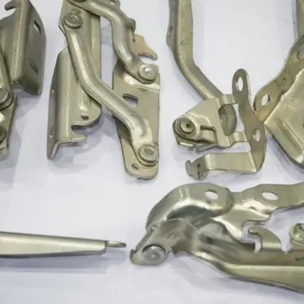 Close-Up Of Various Metallic Mechanical Components, Including Hinges, Levers, And Brackets, Arranged On A Plain White Surface. The Parts Are Made Of A Shiny, Silver Metal And Appear To Be Precision-Engineered For Assembly Using Die Cutting Techniques.