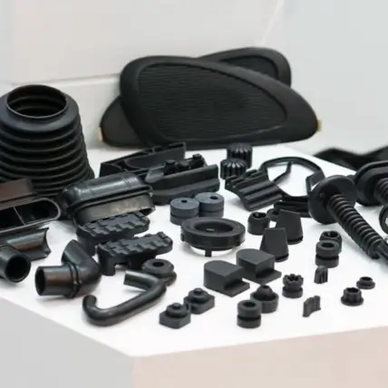 Various Black Plastic Car Parts And Auto Components, Including Grommets, Bushings, And Connectors, Created Through Urethane Casting Service Are Displayed On A White Surface Against A Neutral Background. Some Parts Are Coiled, While Others Have Geometric Shapes Or Complex Designs.