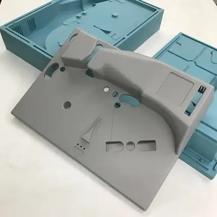 A Photo Of Several Molded Plastic Components Created Utilizing A Urethane Casting Service. The Largest Piece, In The Foreground, Is Gray With Various Cutouts, Holes, And Raised Sections. Behind It, Two Blue Plastic Components Have Compartments And Slots. They Are All Placed On A White Surface.