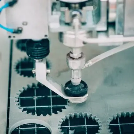 A Close-Up View Of An Industrial Machine Utilizing Waterjet Cutting To Craft Gear-Shaped Pieces From A Sheet Of Material. The Cutting Tool Is Positioned Over The Partially Cut Gears, With Some Already Removed, Showcasing The Precision And Versatility Of This Prototype Technology.