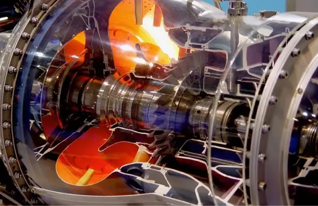 A Close-Up View Of A Jet Engine Turbine Cutaway Model, Showcasing The Internal Components And Structure. The Image Highlights The Engine'S Intricate Parts, Including The Spinning Blades And Various Mechanical Elements, With Some Parts Illuminated In Orange.