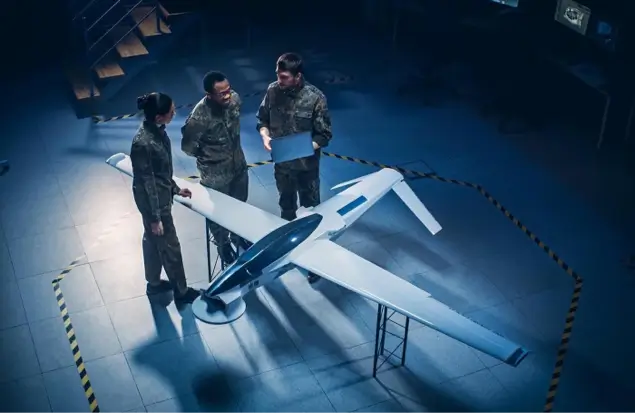 Three Uniformed Military Personnel Stand Around A White Drone Placed On A Table In A Dimly Lit Room. They Are Discussing Something, With One Person Holding A Laptop. The Area Is Marked Off With Yellow And Black Caution Tape.