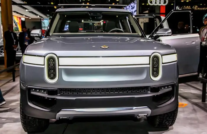 A Silver Electric Suv Is Displayed At An Auto Show. The Vehicle Features A Distinctive Front Design With Vertical Led Headlights, A Prominent Grille, And A Sleek Metallic Finish. The Car Doors Are Open, Showcasing The Interior. Other Vehicles Are Visible In The Background.