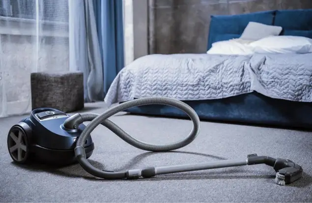 A Black Canister Vacuum Cleaner With A Long Hose And Brush Attachment Rests On A Carpeted Floor In A Bedroom. The Room Features A Neatly Made Bed With Blue And White Bedding, Sheer White Curtains, And A Modern Gray Ottoman.