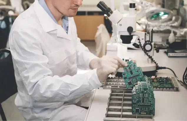 A Person Wearing A White Lab Coat And Gloves Inspects Green Circuit Boards At A Workstation With A Microscope. The Station Has Various Electronic Components And Tools Organized Around It.