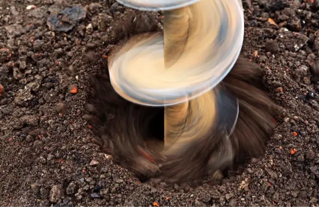 A Closeup Of A Drill Bit Rapidly Spinning And Digging Into The Soil, Creating A Hole. The Ground Around The Hole Appears Dark And Slightly Rocky. The Motion Blur Emphasizes The Speed Of The Drilling.