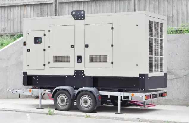 A Large, White Industrial Generator Mounted On A Trailer With Two Wheels Is Positioned Outdoors Near A Concrete Wall. The Generator Features Multiple Vents And Access Panels. The Trailer Has A Sturdy Frame And Hitch For Towing.