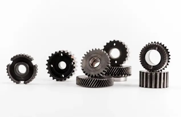 A Group Of Metallic Gears Of Varying Sizes And Designs Are Arranged Against A White Background. The Gears Are Standing Upright, Lying Flat, Or Leaning Against One Another. The Image Showcases Their Intricate Teeth And Mechanical Details.