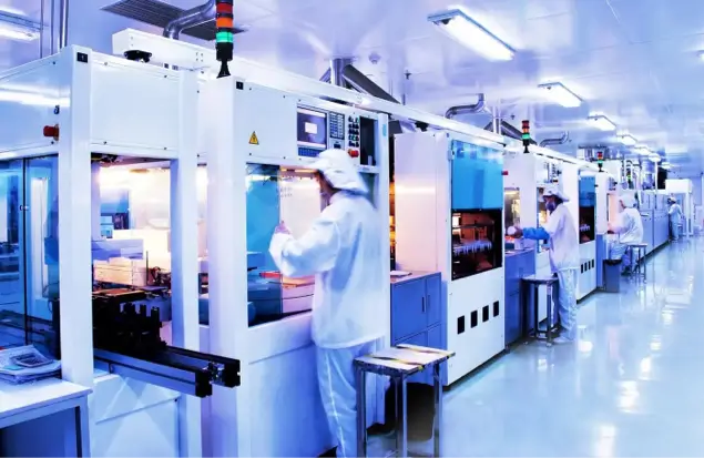Workers In White Cleanroom Suits Operate Machinery In A Brightly Lit, Modern Factory Or Laboratory. The Environment Is Meticulously Clean, With Several Workstations And Equipment Lined Up Against The Walls, Indicating A High-Tech Production Process.