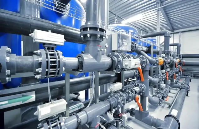 The Image Shows A Complex Setup Of Industrial Pipes And Valves Inside A Facility With A Metallic Interior. Various Types Of Pipes, Gauges, And Control Systems Are Neatly Organized, Indicating A Highly Technical Environment Likely Related To Water Or Gas Processing.