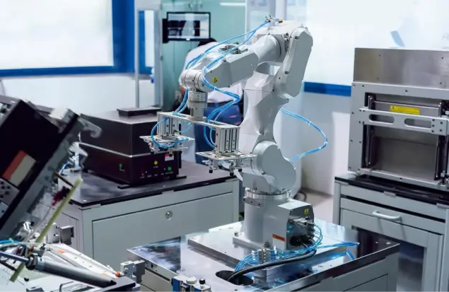 A Robotic Arm Equipped With Pneumatic Tubes Operates In A High-Tech Laboratory. Surrounding It Are Various Pieces Of Equipment, Including A Monitor And Other Machinery. The Setup Appears Designed For Precision Tasks, Possibly In Manufacturing Or Research.