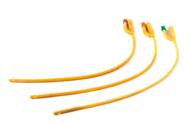Three Yellow Foley Catheters With Different Colored Tips (Red, Green, And Blue) Are Lined Up Diagonally Across A White Background. Each Catheter Ends With A Balloon Port And A Drainage Port.