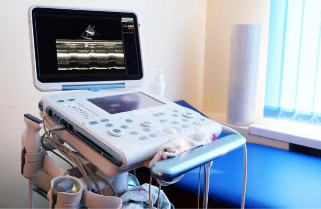 A Medical Ultrasound Machine With A Screen Displaying A Scan Image Sits On A Cart. The Machine Has Various Buttons And Knobs For Control. A Roll Of Paper Towels And A Bottle Of Hand Sanitizer Are Nearby. A Blue Cushioned Exam Table Is In The Background.